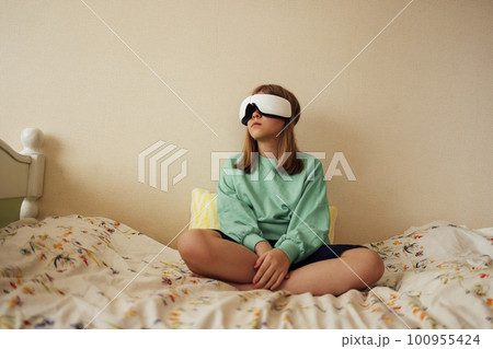 Pretty female teenager is sitting in VR glasses on bed in her bedroom. 100955424
