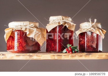 Jars of lingonberry and pear jam with craft paper lids on wooden shelf next to fresh lingonberries on brown background Jars of lingonberry and pear jam with craft paper lids on wooden shelf next to fresh lingonberries on brown background 100958770