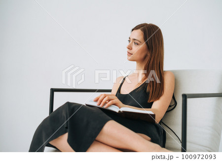 Beautiful young woman with dark long hair in black dress reading book on chair in white interior Beautiful young woman with dark long hair in black dress reading book on chair in white interior 100972093