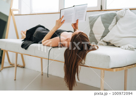 Beautiful young woman with dark long hair in black dress reading book in white interior 100972094