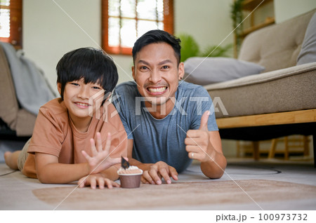 Happy Asian dad showing thumb up while laying on the living room floor with his son 100973732