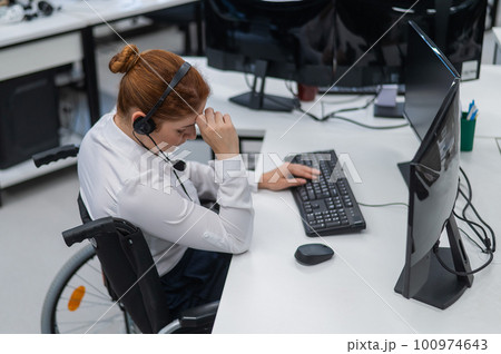 Caucasian woman in a wheelchair sits at her desk and holds her head. Female call center worker. 100974643