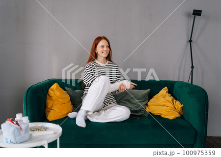 Portrait of cheerful young woman with broken hand wrapped in white plaster bandage sitting on sofa in living room smiling looking away. Positive redhead female with broken arm from accident at home. Portrait of cheerful young woman with broken hand wrapped in white plaster bandage sitting on sofa in living room smiling looking away. Positive redhead female with broken arm from accident at home. 100975359