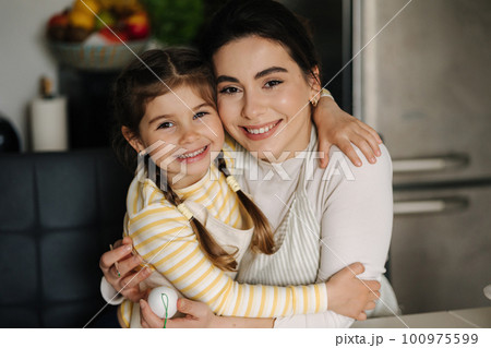 Close-up portrait of daughter with mom on kitchen. Beautiful girls dressed in apron Close-up portrait of daughter with mom on kitchen. Beautiful girls dressed in apron 100975599