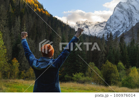 man stands against the backdrop of a mountain landscape with his hands raised high. Autumn or summer, in the foreground a dense green forest, behind it are snow-capped mountains, a joyful tourist man stands against the backdrop of a mountain landscape with his hands raised high. Autumn or summer, in the foreground a dense green forest, behind it are snow-capped mountains, a joyful tourist 100976014