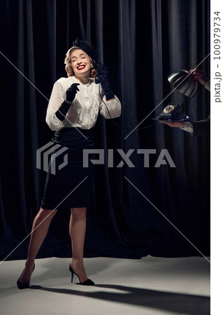 Adorable woman wearing vintage clothes holding phone and talking with smiling over dark background 100979734