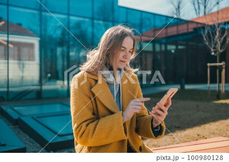 Photo portrait of blonde happy student woman 20s holding phone wearing a yellow coat and sitting on bench in city spring park outdoors resting use mobile cell phone chat online. 100980128