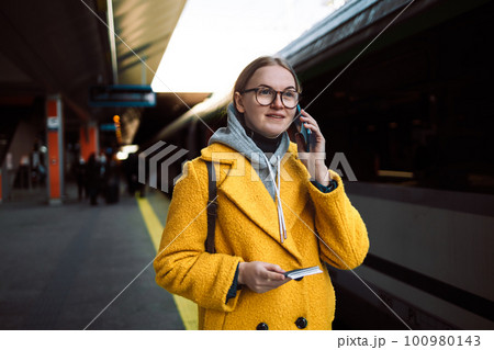 30s woman makes telephone call in roaming wears casual sweatshirt and yellow coat had glad expression waiting on a train station platform with passport and tickets enjoys smartphone talking. 30s woman makes telephone call in roaming wears casual sweatshirt and yellow coat had glad expression waiting on a train station platform with passport and tickets enjoys smartphone talking. 100980143
