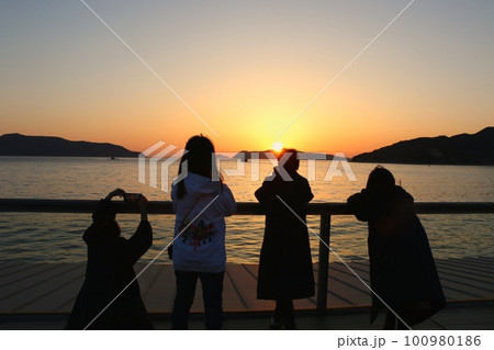 海と山に沈む夕陽を眺める人達 100980186