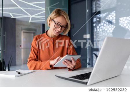 Young successful businesswoman at workplace is satisfied with results of achievement at work smiling and using tablet computer, blonde programmer is testing new software in application. 100980280