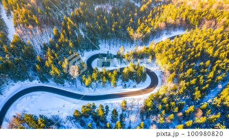 Asphalt road serpentine in snowy wintertime. Cold winter and sunny day above forest road with illuminated trees by rising sun. Aerial view from drone. Asphalt road serpentine in snowy wintertime. Cold winter and sunny day above forest road with illuminated trees by rising sun. Aerial view from drone. 100980820