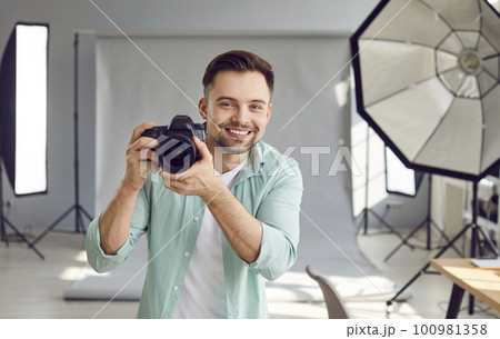 Happy smiling photographer holding a camera standing in professionally studio with light equipment. Happy smiling photographer holding a camera standing in professionally studio with light equipment. 100981358