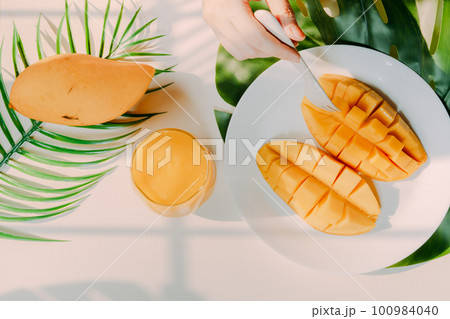 top view and flat lay summer fruit from beauty woman hand hold mango juice and use fork for eat mango slice with shadow of tropical leaf and white background 100984040