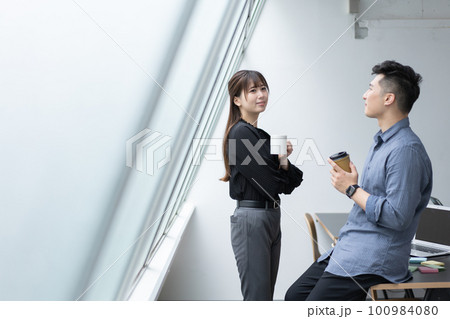 オフィスで飲み物を手に立って会話する若い男女 100984080