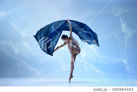 Adorable dancer wearing beige bodysuit dancing with fabric silk over blue background Adorable dancer wearing beige bodysuit dancing with fabric silk over blue background 100984392