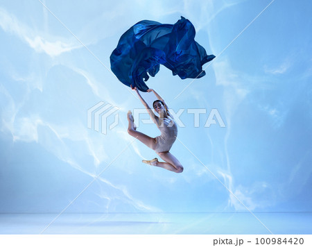 Shot of flexible ballerina, dancer in high jump with navy fabric over blue background. Action, motion, art concept Shot of flexible ballerina, dancer in high jump with navy fabric over blue background. Action, motion, art concept 100984420
