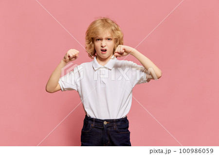 Little boy, child emotively posing in white shirt, showing fists and little anger, posing against pink studio background. Childhood and emotions Little boy, child emotively posing in white shirt, showing fists and little anger, posing against pink studio background. Childhood and emotions 100986059