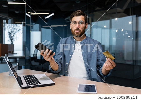 Portrait of disappointed and deceived businessman inside office, mature man looking at camera confused holding bank credit card and phone, employee got rejection error. Portrait of disappointed and deceived businessman inside office, mature man looking at camera confused holding bank credit card and phone, employee got rejection error. 100986211