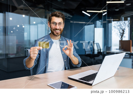 Successful businessman inside office building at workplace smiling and looking at camera, mature adult man holding and showing bank credit card, smiling contentedly. Successful businessman inside office building at workplace smiling and looking at camera, mature adult man holding and showing bank credit card, smiling contentedly. 100986213