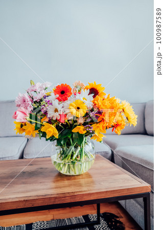 Vase with huge multicolor various flower bouquet on the coffee table with blurred background of modern cozy light living room with gray couch sofa. Giving flowers. Mother's day, birthday gift. 100987589