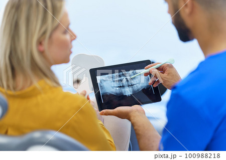 Rear view of dentist showing x-ray scan at digital tablet to his patient. 100988218