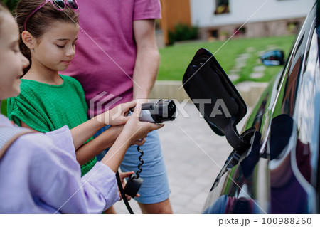 Close-up of father holding power supply cable with his little daughters and charging their electric car. Close-up of father holding power supply cable with his little daughters and charging their electric car. 100988260