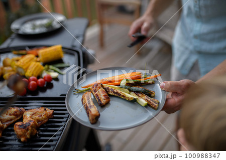 Father putting grilled meat and vegetable on plate to his son during family summer garden party. 100988347