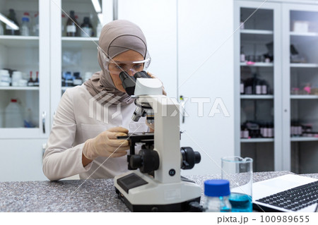 A young Muslim woman is a laboratory assistant, a doctor sitting at a table in a medical diagnostic center. Examines tests, looks into a microscope. A young Muslim woman is a laboratory assistant, a doctor sitting at a table in a medical diagnostic center. Examines tests, looks into a microscope. 100989655