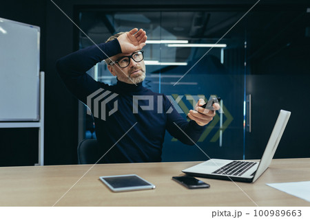 An elderly man, a businessman, sits in the office at his desk and turns on the air conditioner with the remote control. Suffers from heat, suffocation, holds his head, wipes sweat. An elderly man, a businessman, sits in the office at his desk and turns on the air conditioner with the remote control. Suffers from heat, suffocation, holds his head, wipes sweat. 100989663
