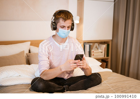 guy in headphones on the bed in a protective mask with a phone in his hand guy in headphones on the bed in a protective mask with a phone in his hand 100991410