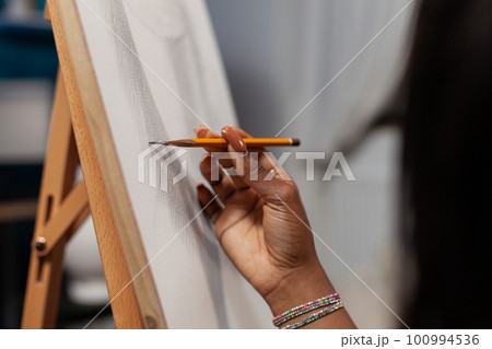 Close up of hand of unrecognizable african american female artist making strokes with pencil on canvas easel in drawing workshop. Unidentified creative person finishing artwork at studio. 100994536