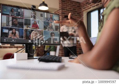 African american woman gesturing in internet meeting with colleagues while roommate is working on laptop in home living room. Startup employee in internet call with diverse team using pc and webcam. African american woman gesturing in internet meeting with colleagues while roommate is working on laptop in home living room. Startup employee in internet call with diverse team using pc and webcam. 100994597