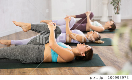 Class of young sporty women stretching gluteal muscle while lying on back during yoga Pilates workout on mats in gym studio 100998978