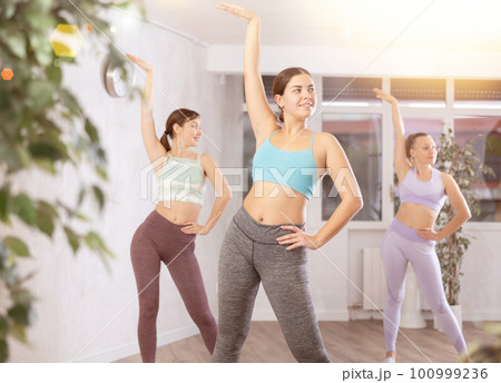 Group of young energetic women in tight activewear warming up with dancing elements before training together in fitness center Group of young energetic women in tight activewear warming up with dancing elements before training together in fitness center 100999236