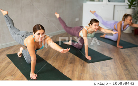 Diligent women practicing balancing table pose of yoga in light fitness room Diligent women practicing balancing table pose of yoga in light fitness room 100999442