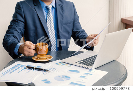 Businessman hand holding coffee cup, analysis the graph with laptop at the home office for setting challenging business goals And planning to achieve the new target 100999778