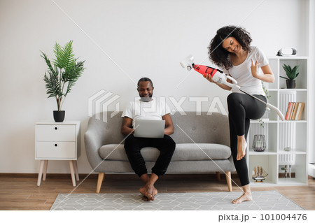 Woman dancing with cordless vacuum while man using laptop Woman dancing with cordless vacuum while man using laptop 101004476
