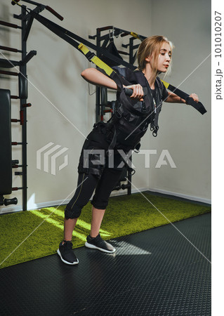 Beautiful blonde woman wearing in sensors and belts, fastens with velcro is practicing EMS fitness in a gym performing traction exercises. Beautiful blonde woman wearing in sensors and belts, fastens with velcro is practicing EMS fitness in a gym performing traction exercises. 101010207