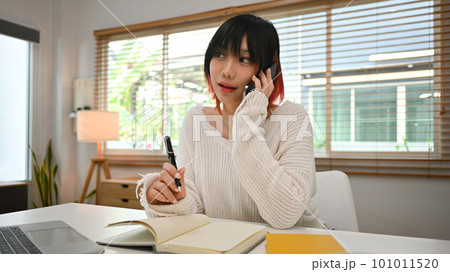 Charming young Asian woman freelancer having phone conversation and making note on notebook Charming young Asian woman freelancer having phone conversation and making note on notebook 101011520