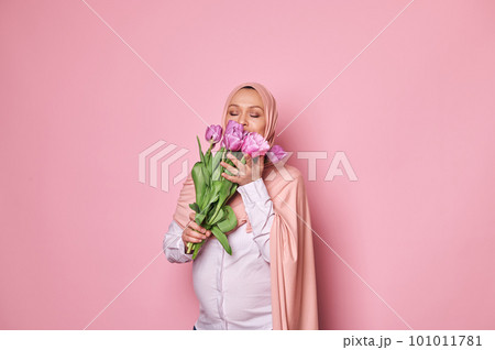 Waist-up portrait of a delightful beautiful Muslim woman in hijab, sniffing a cute bouquet of purple tulips, on isolated pink background. The concept of Women's Day, maternity, pregnancy, Mother's day Waist-up portrait of a delightful beautiful Muslim woman in hijab, sniffing a cute bouquet of purple tulips, on isolated pink background. The concept of Women's Day, maternity, pregnancy, Mother's day 101011781