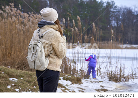 Mother watching a child playing on the shore of a frozen lake in a winter park 101012471