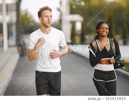 African american fitness model and caucasian man jogging outdoors. Woman wearing black sportswear. Man wearing white t-shirt. African american fitness model and caucasian man jogging outdoors. Woman wearing black sportswear. Man wearing white t-shirt. 101013853