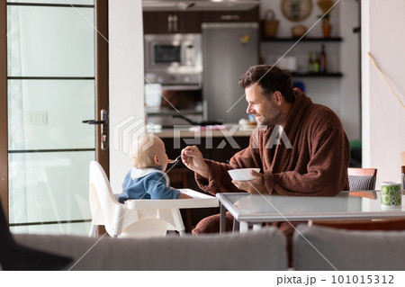 Father wearing bathrope spoon feeding hir infant baby boy child sitting in high chair at the dining table in kitchen at home in the morning. 101015312