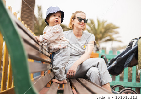 Young mother with her cute infant baby boy child on bench on urban children's playground on warm summer day 101015313