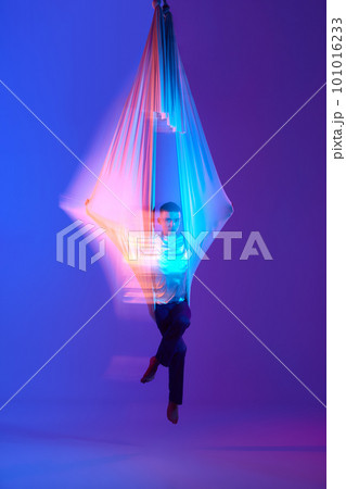 Young man, aerial acrobat, gymnast training with aerial fabric against gradient blue purple background in neon with mixed lights 101016233