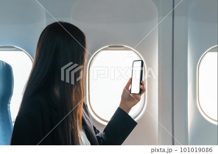 A female traveler passenger sits at the window seat in economy class, using his smartphone, holding a mobile phone white screen mockup. close-up image 101019086