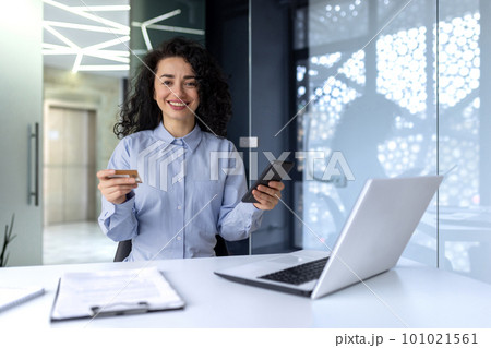 A young Muslim woman makes online purchases through a phone application, pays with a credit card. Sitting in the office at the table with a laptop, looking at the camera. A young Muslim woman makes online purchases through a phone application, pays with a credit card. Sitting in the office at the table with a laptop, looking at the camera. 101021561