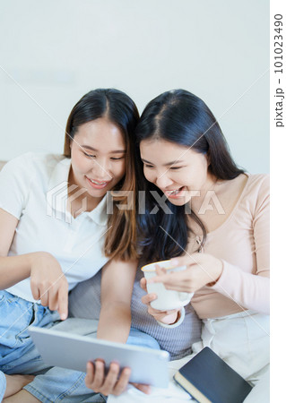 lgbtq, lgbt concept, homosexuality, portrait of two asian women posing happy together and loving each other while playing tablet at sofa. lgbtq, lgbt concept, homosexuality, portrait of two asian women posing happy together and loving each other while playing tablet at sofa. 101023490