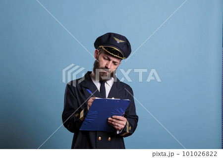 Airplane pilot holding clipboard, writing, concentrated aviator with documents. Aircraft captain in professional uniform filling form in airport front view, studio medium shot on blue background Airplane pilot holding clipboard, writing, concentrated aviator with documents. Aircraft captain in professional uniform filling form in airport front view, studio medium shot on blue background 101026822