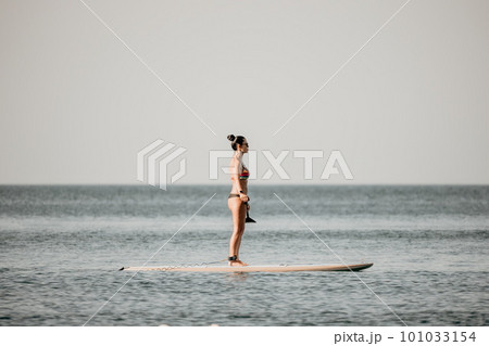 Sea woman sup. Silhouette of happy middle aged tanned woman in rainbow bikini, surfing on SUP board, confident paddling through water surface. Idyllic sunset. Active lifestyle at sea or river. 101033154